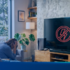 Pessoa sentada no sofá com TV exibindo símbolo de energia bloqueada — representa oscilações elétricas